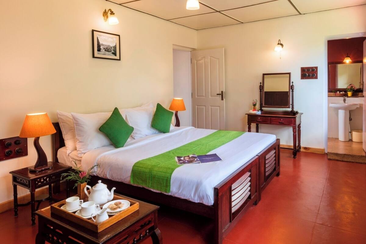 Une chambre d’hôtel bien rangée, avec un lit, des tables de chevet, une commode, un secrétaire, un miroir et une salle de bain visible à travers une porte ouverte.