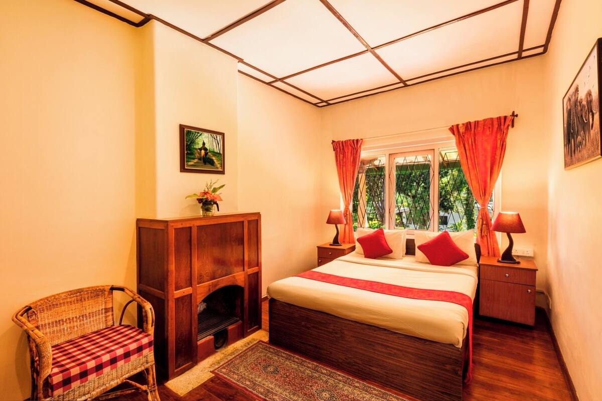 Une chambre à coucher avec un lit en bois, une cheminée, un fauteuil en osier et une fenêtre avec des rideaux.