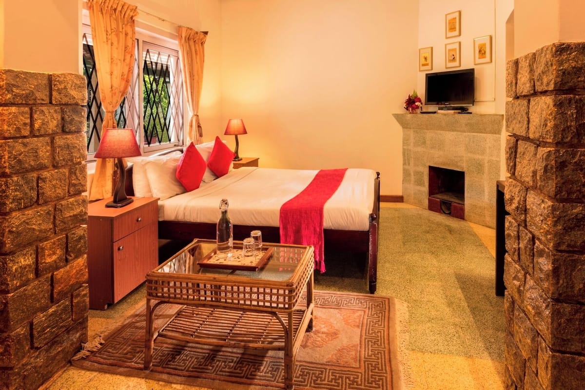 Une chambre confortable avec une cheminée en pierre, un lit avec des oreillers rouges, une table de chevet avec des lampes et une petite table avec une bouteille et des verres.