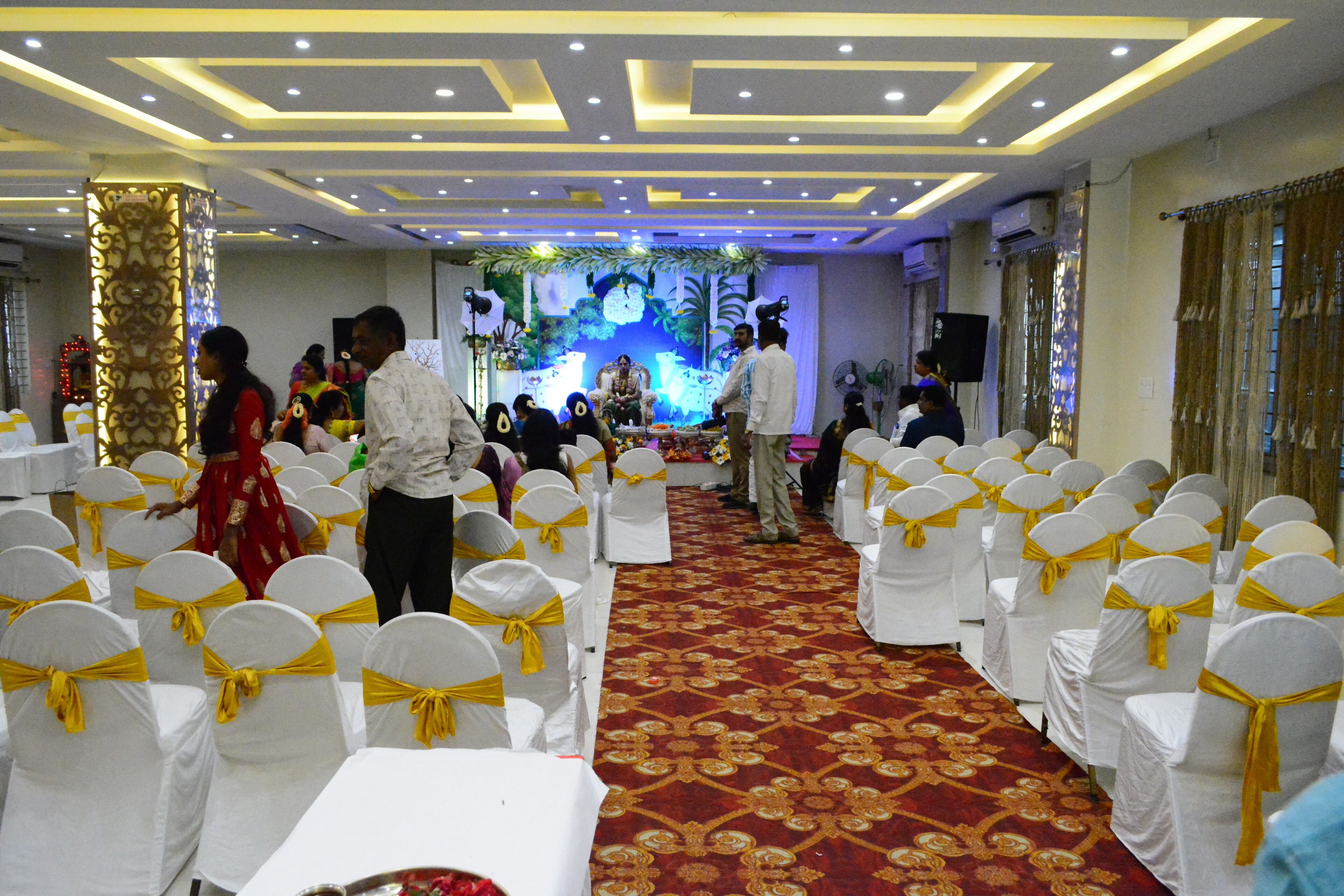 A banquet hall with rows of white chairs with yellow sashes, a red patterned carpet, and a stage with floral decorations.