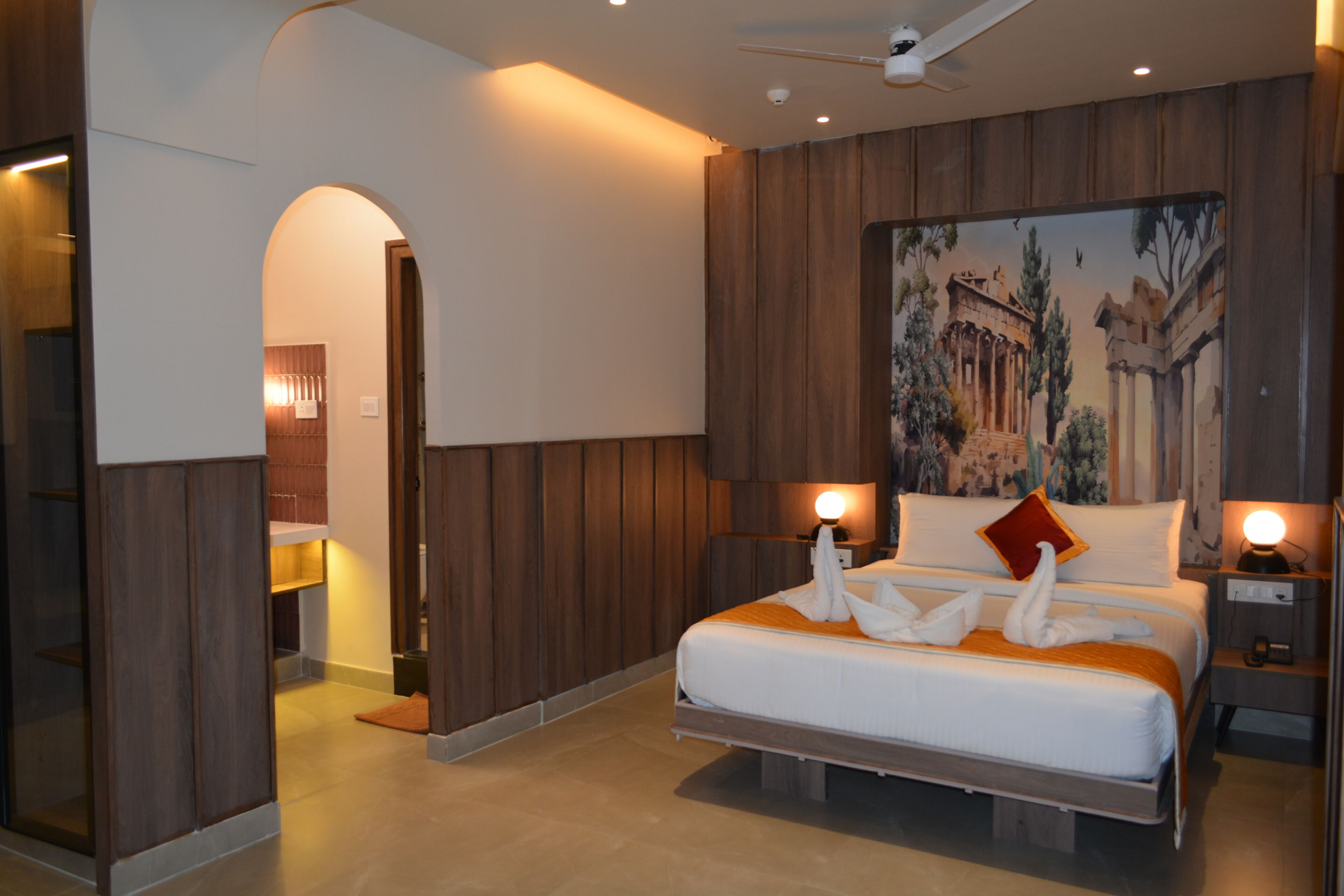A modern hotel room with a large bed, wooden paneling, a painting of ancient ruins, and a ceiling fan.