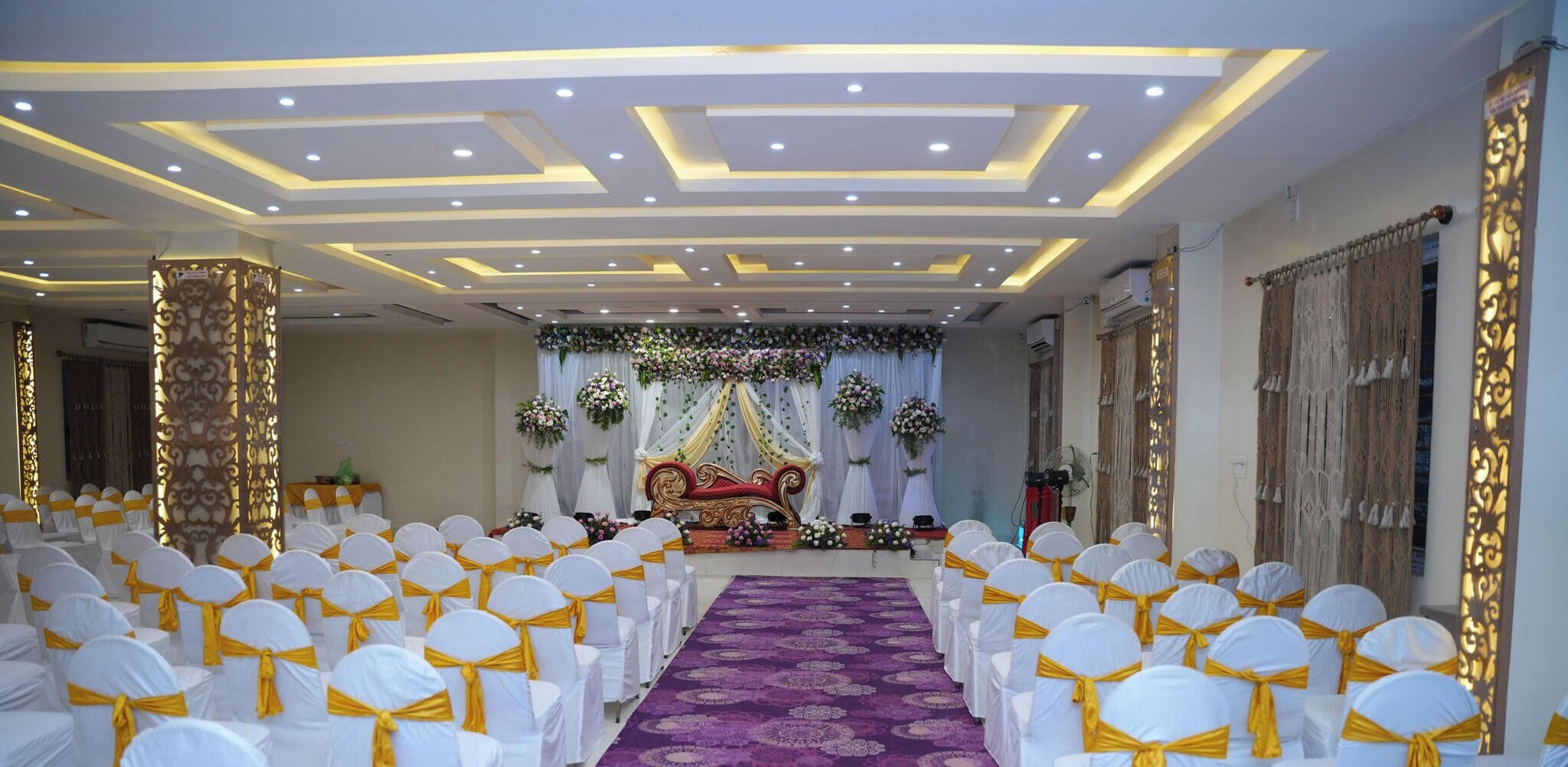 A banquet hall with rows of white chairs with yellow sashes, a purple carpet, and a stage with floral decorations.