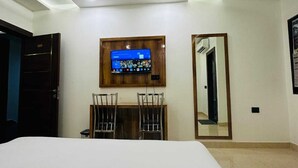 A hotel room with a bed, a desk with two chairs, a flat-screen TV mounted on the wall, a ceiling fan, and a mirror on the wall. - Chandan Palace (Agra)