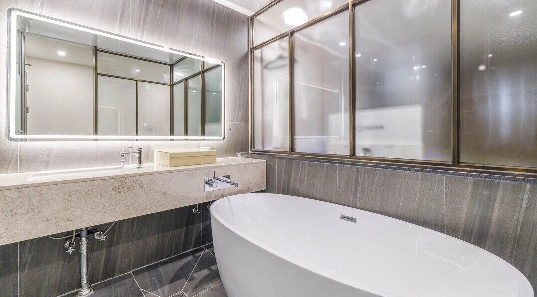A modern bathroom with a freestanding bathtub, a large mirror with integrated lighting, and a window with frosted glass.