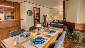 A dining area with a wooden table set for four, a stone fireplace, and a living room with a sofa and a ladder shelf.