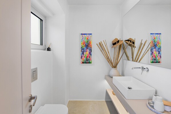 A modern bathroom with a white sink, mirror, and a window. There are two abstract paintings on the walls and a vase with reeds.