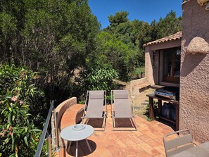 A patio with two lounge chairs and a small table, surrounded by greenery and a stone building.