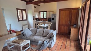 Ein gemütliches Wohnzimmer mit einer Couch, einem kleinen Tisch und einem Sessel. Im Hintergrund ist eine Küchenzeile mit Schränken und einem Kühlschrank zu sehen.