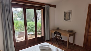Ein Zimmer mit einem Bett, einem Schreibtisch und einem Stuhl. Ein Fenster mit Vorhängen bietet Blick ins Grüne.
