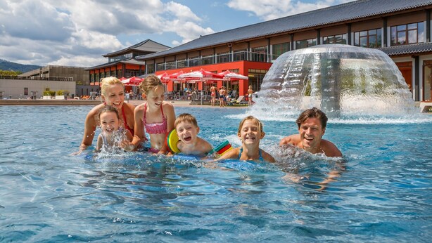 A swimming pool with people enjoying the water, a building with red umbrellas in the background, and a waterfall feature. - Superior Apartment with IR-Sauna (No Deposit) (Eisenerz)