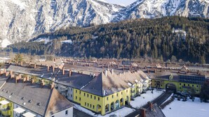 A town with snow-covered mountains in the background and a row of yellow buildings with multiple stories and visible chimneys. - Superior Apartment with IR-Sauna (No Deposit) (Eisenerz)