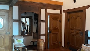 Een ruime kamer met hoog plafond, houten balken, een kroonluchter, een rieten stoel, een beklede bank en een deur met spiegel.