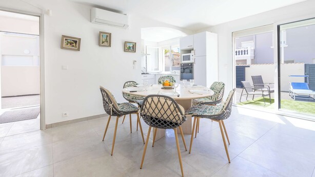 A modern dining area with a round table, six patterned chairs, a white ceiling, and a view of a garden through glass doors. - Apartment Near the Sea – Valras-Plage (Valras-Plage)