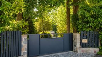 A gated entrance with a stone wall and a metal gate, surrounded by lush green trees.