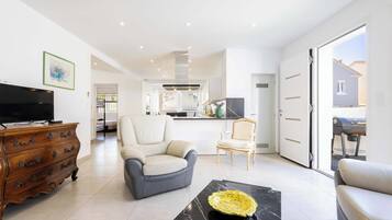 Un salon moderne avec un téléviseur, une commode en bois, deux fauteuils, une table basse en marbre et une cuisine en arrière-plan.