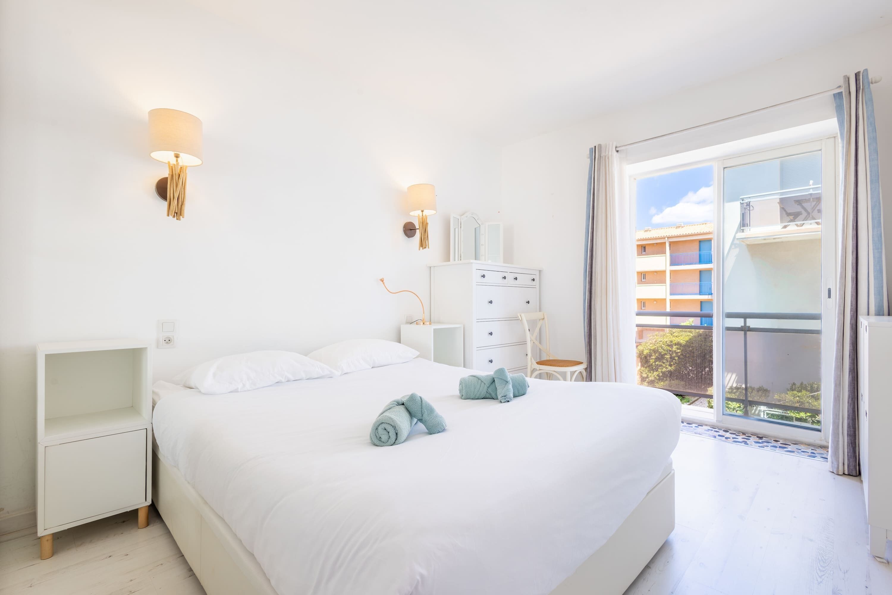 Une chambre à coucher comprenant un lit, une table de chevet, une commode et donnant sur l’extérieur.