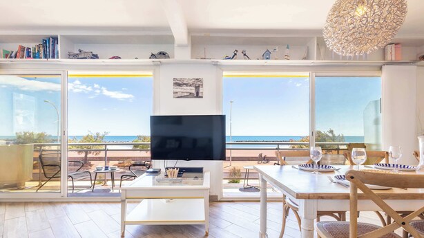 A modern living room with a TV, dining table, chairs, and large windows overlooking the sea.