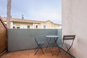 Een klein buitenterras met een ronde tafel en twee stoelen, een betegelde vloer, en een gebouw op de achtergrond.