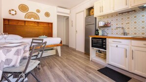A compact room with a bed, a dining table with chairs, a kitchenette with a refrigerator, oven, and microwave, and a wall-mounted air conditioner. - Valras-Loc – Studio with terrace (Valras-Plage)