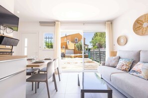 A modern living room with a sofa, a dining area with chairs and a table, and a view of a courtyard with a building in the background. - Private Garden Studio, Steps from Valras-Plage (Valras-Plage)