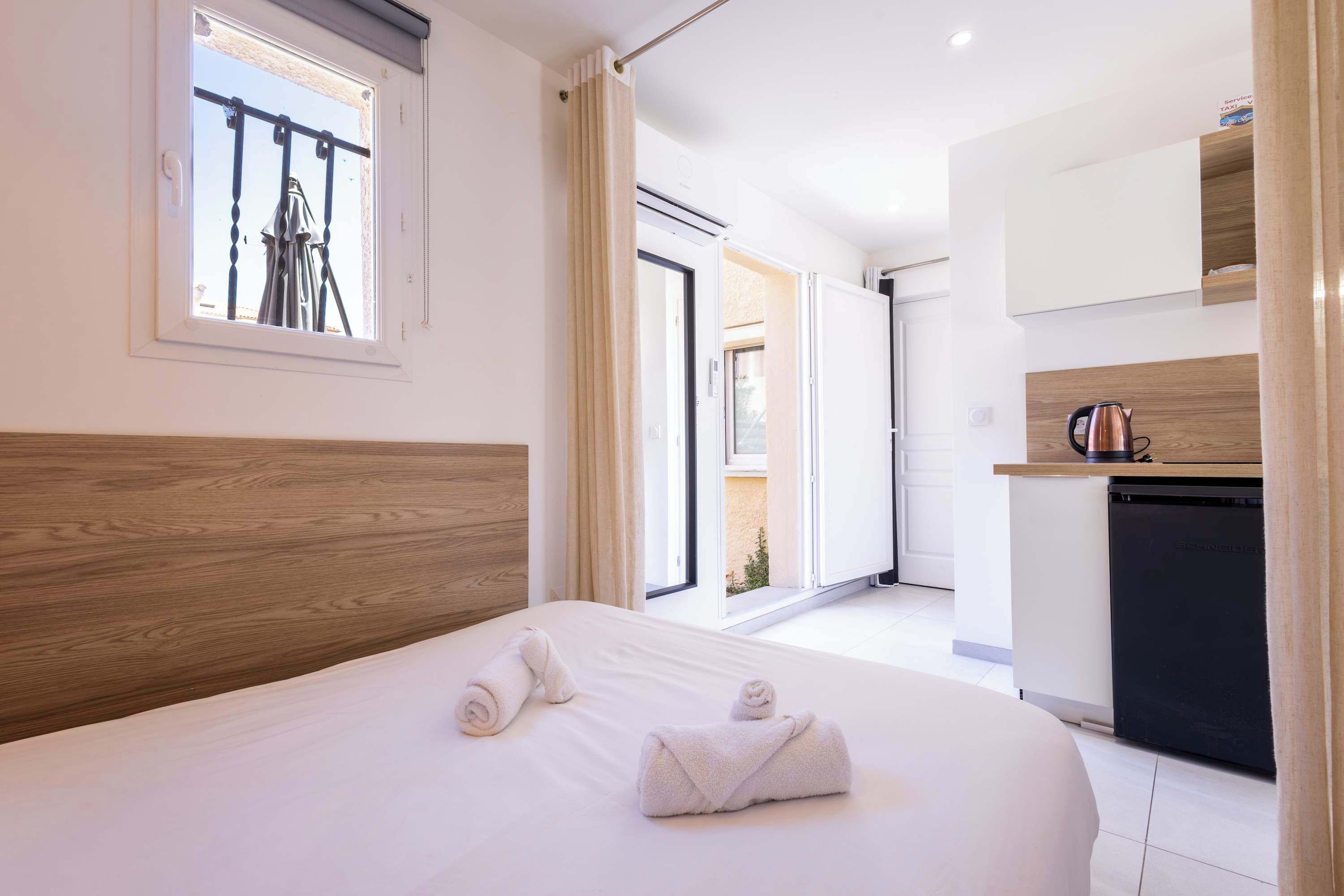 A hotel room with a wooden headboard, a bed with white linens, a window with a view of the outside, and a kitchenette with a coffee maker.