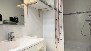 Une salle de bain avec un lavabo, des toilettes et une douche, le tout de couleur blanche. Il y a des étagères au-dessus du lavabo et un miroir au mur.