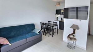 A living room with a blue sofa, dining table, chairs, a small side table with a lamp, and a kitchen area in the background. - Loggia Verde – Charming studio 450 m from the sea (Sérignan)