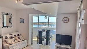 A living room with a sofa, TV, and a balcony with a view. - Comfortable Studio with Harbour View (Valras-Plage)