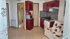 A compact kitchen with red cabinets, a microwave, and a refrigerator. There is a small seating area with a patterned sofa. - Comfortable Studio with Harbour View (Valras-Plage)