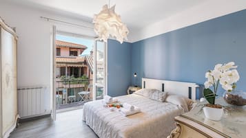 Une chambre à coucher comprenant un lit, une table de chevet, une commode, un luminaire suspendu, une fenêtre donnant sur des immeubles et un balcon avec des chaises et des plantes.