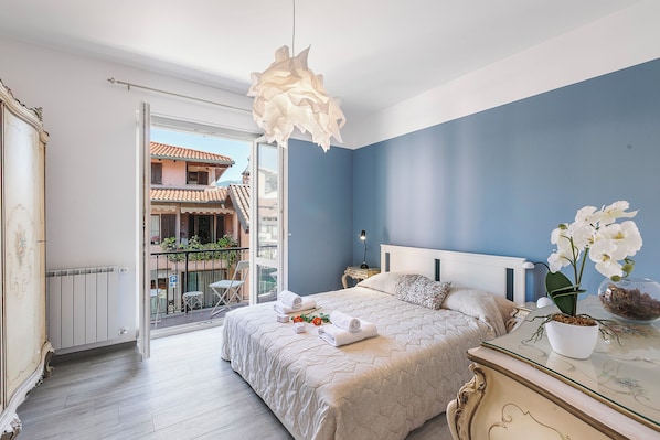 A bedroom with a bed, a nightstand, a dresser, a hanging light fixture, a window with a view of buildings, and a balcony with chairs and plants.