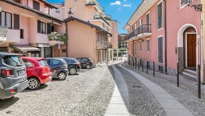 A narrow cobblestone street lined with pastel-colored buildings, parked cars, and a clear blue sky. - Incanto Del Lago 50m to Lake Maggiore (Baveno)