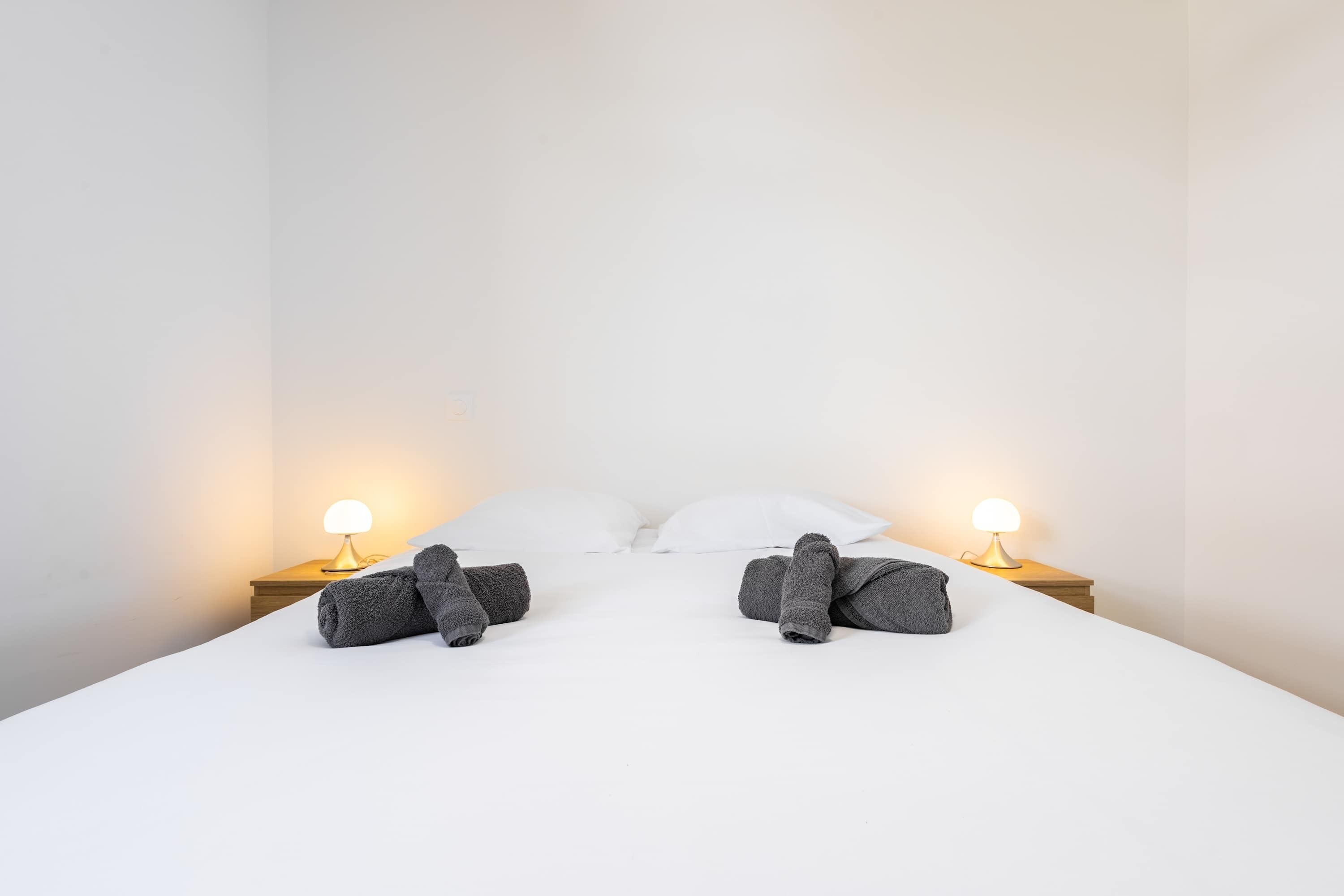 Una cama bien tendida con dos almohadas y dos toallas enrolladas, con lámparas de noche a cada lado sobre mesitas de madera.