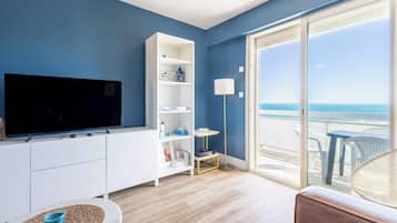 A living room with a TV, white shelving, a small table with a blue bowl, and a view of the sea through a glass door.