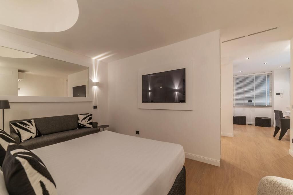 A modern bedroom with a large bed, a mirror, a wall-mounted artwork, and a view into a connected room.