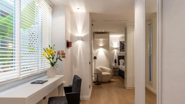 A modern hotel room with a wooden floor, a desk with a chair, a bedside table, and a large window with blinds. - JOIVY Elegant Suites Near Villa Borghese (Rome)