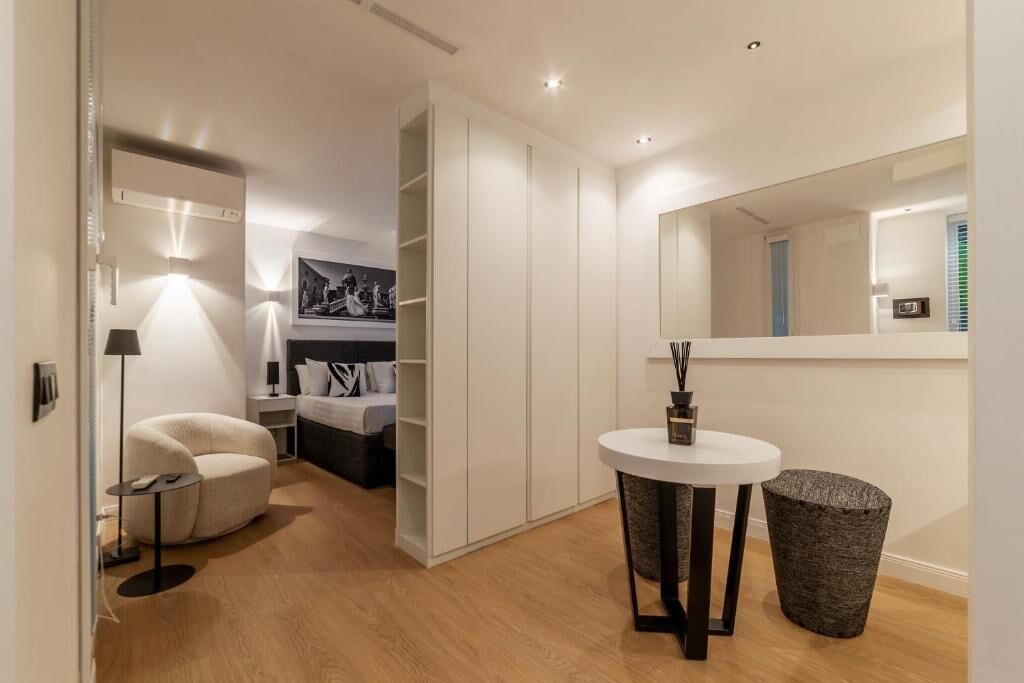 A modern hotel room with a round white table, a black trash bin, a white sofa, and a large mirror.
