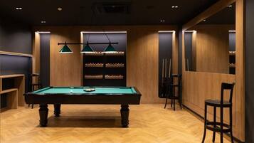 A billiards room with a pool table, green hanging lamps, and shelves with billiard balls.