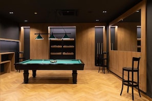 Una sala de billar con mesa de pool, lámparas colgantes verdes y estantes con bolas de billar.