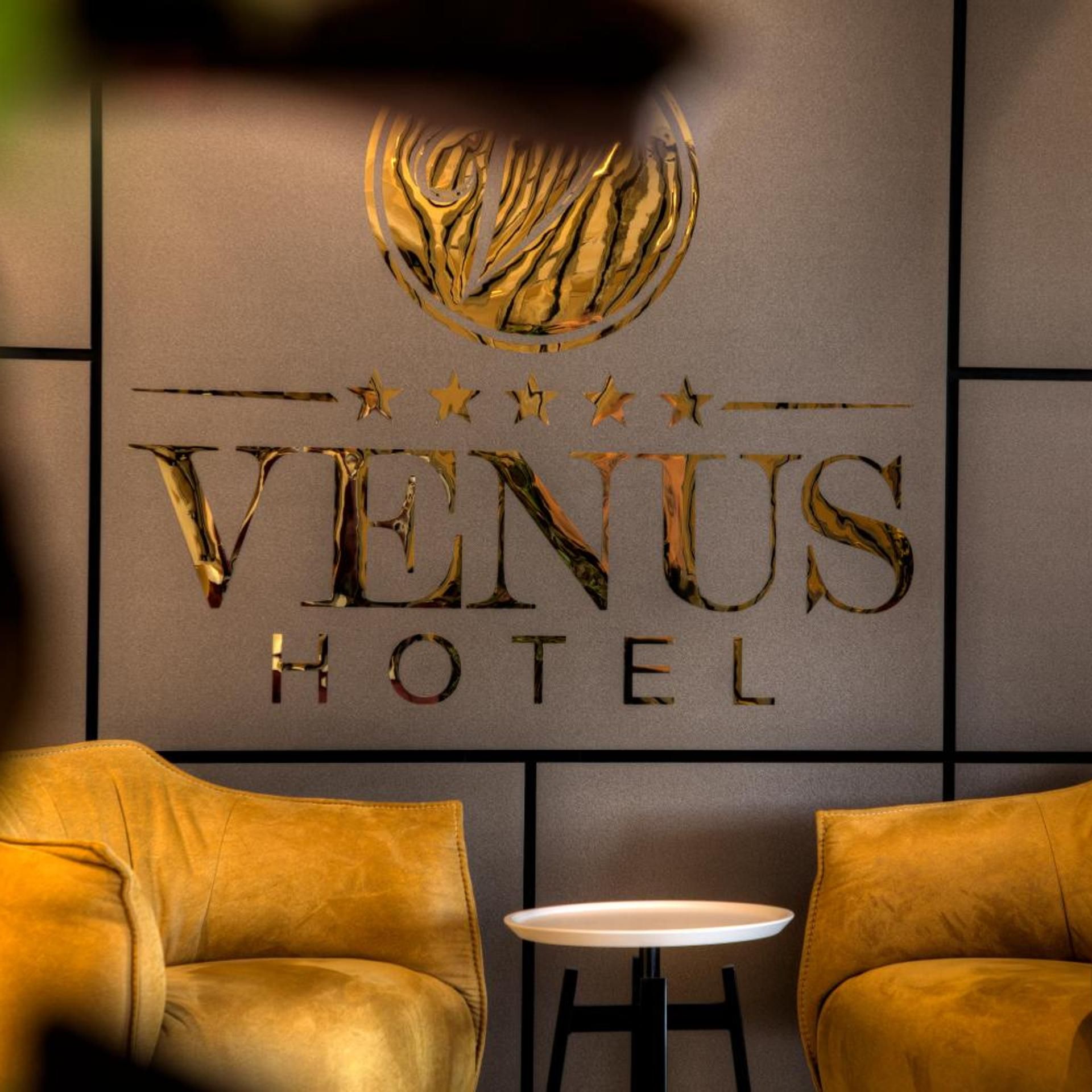 A hotel sign with the name 'VENUS HOTEL' and a decorative emblem above it. There are two chairs and a small round table in front of the sign.