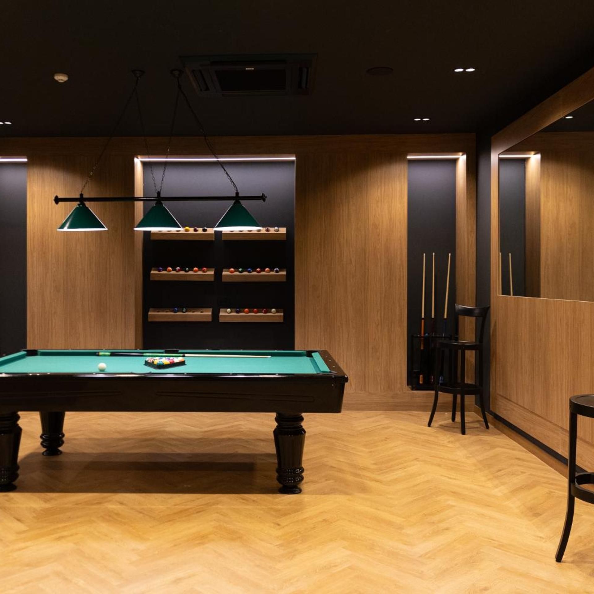 Una mesa de billar con superficie de fieltro verde, rodeada de paredes de madera y un suelo con diseño de espiga.