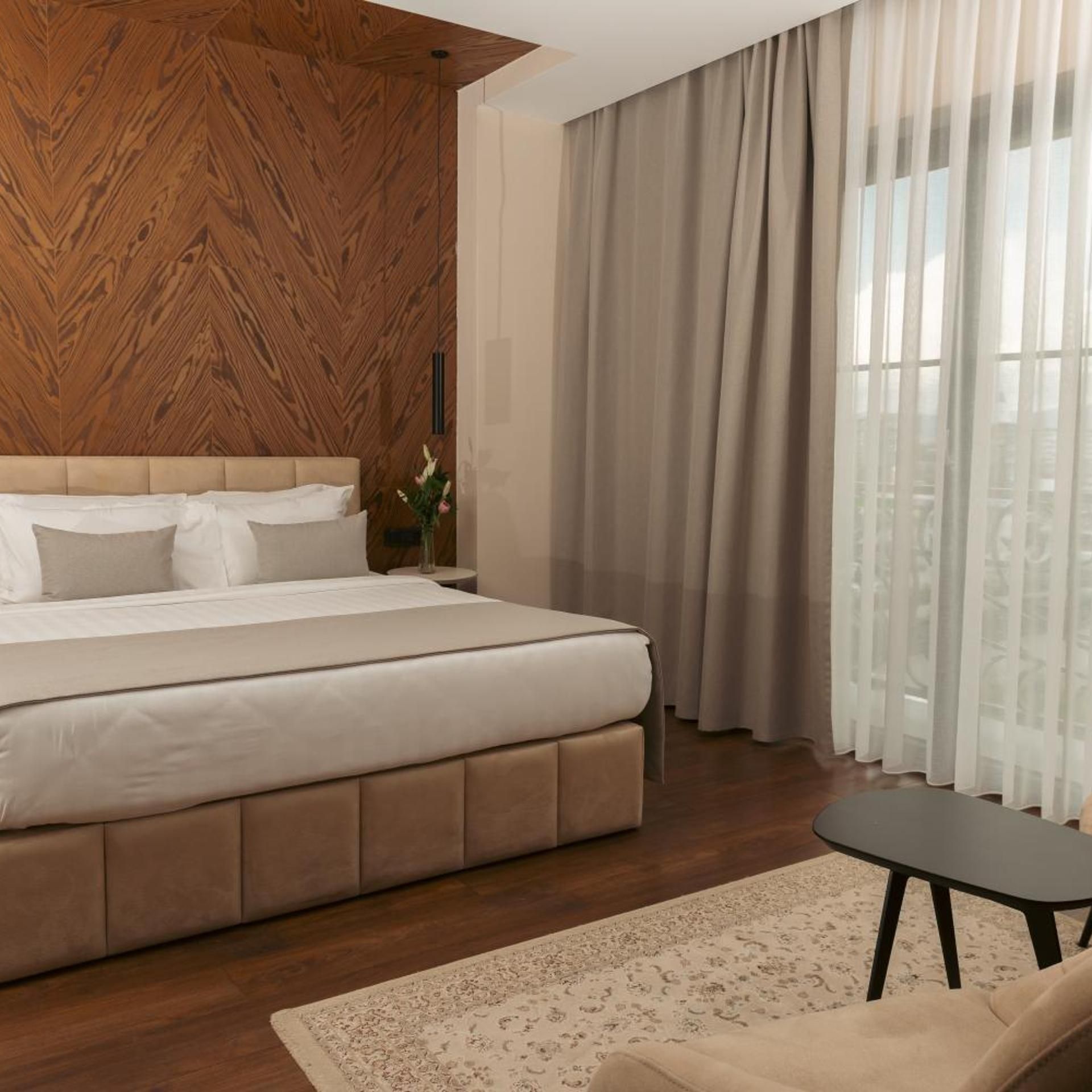 A modern bedroom with a wooden accent wall, a large bed, a small table, and a view through sheer curtains.