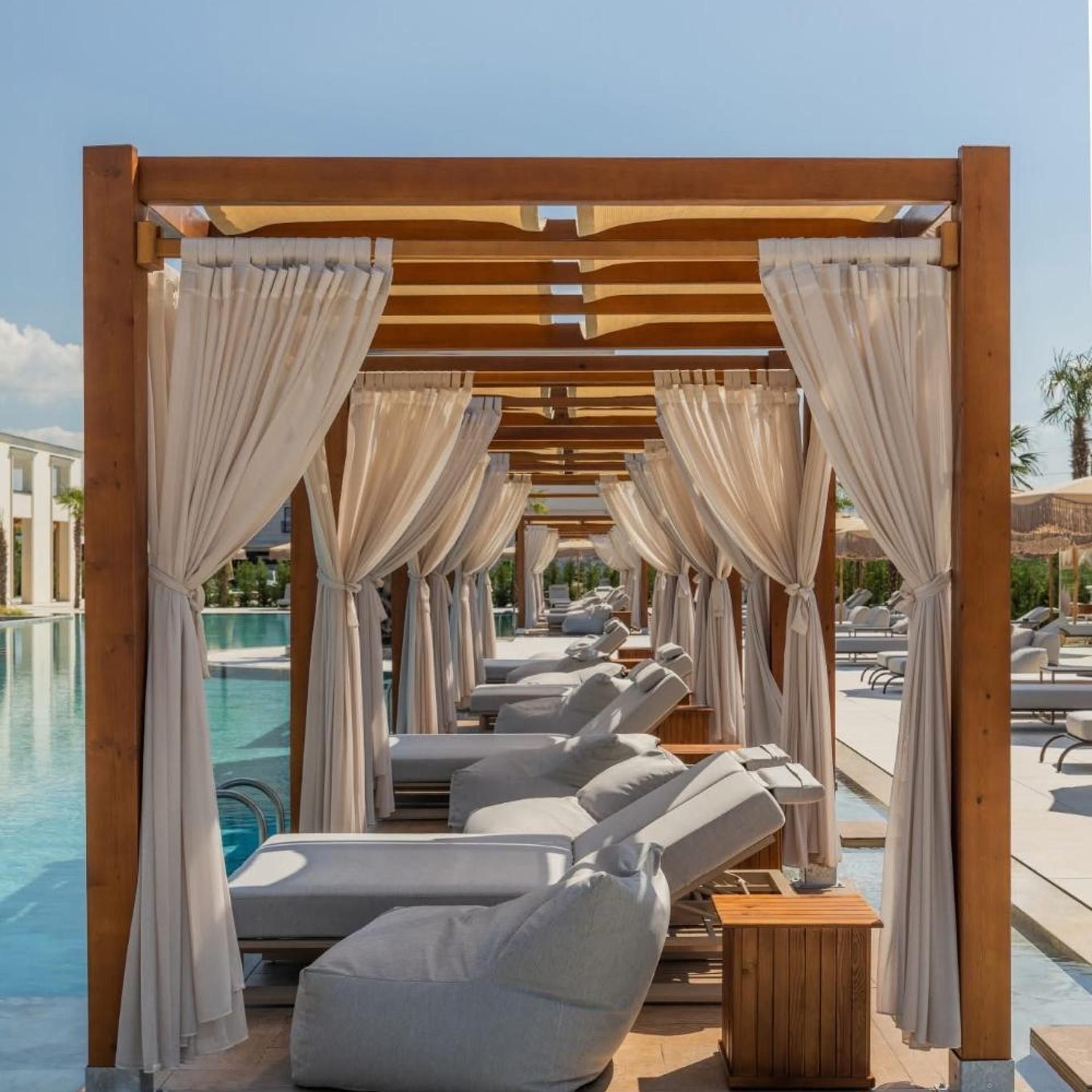 Zona de piscina con tumbonas bajo una pérgola de madera con cortinas colgantes.