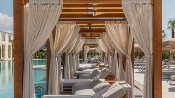 Zona de piscina con tumbonas bajo una pérgola de madera con cortinas colgantes.