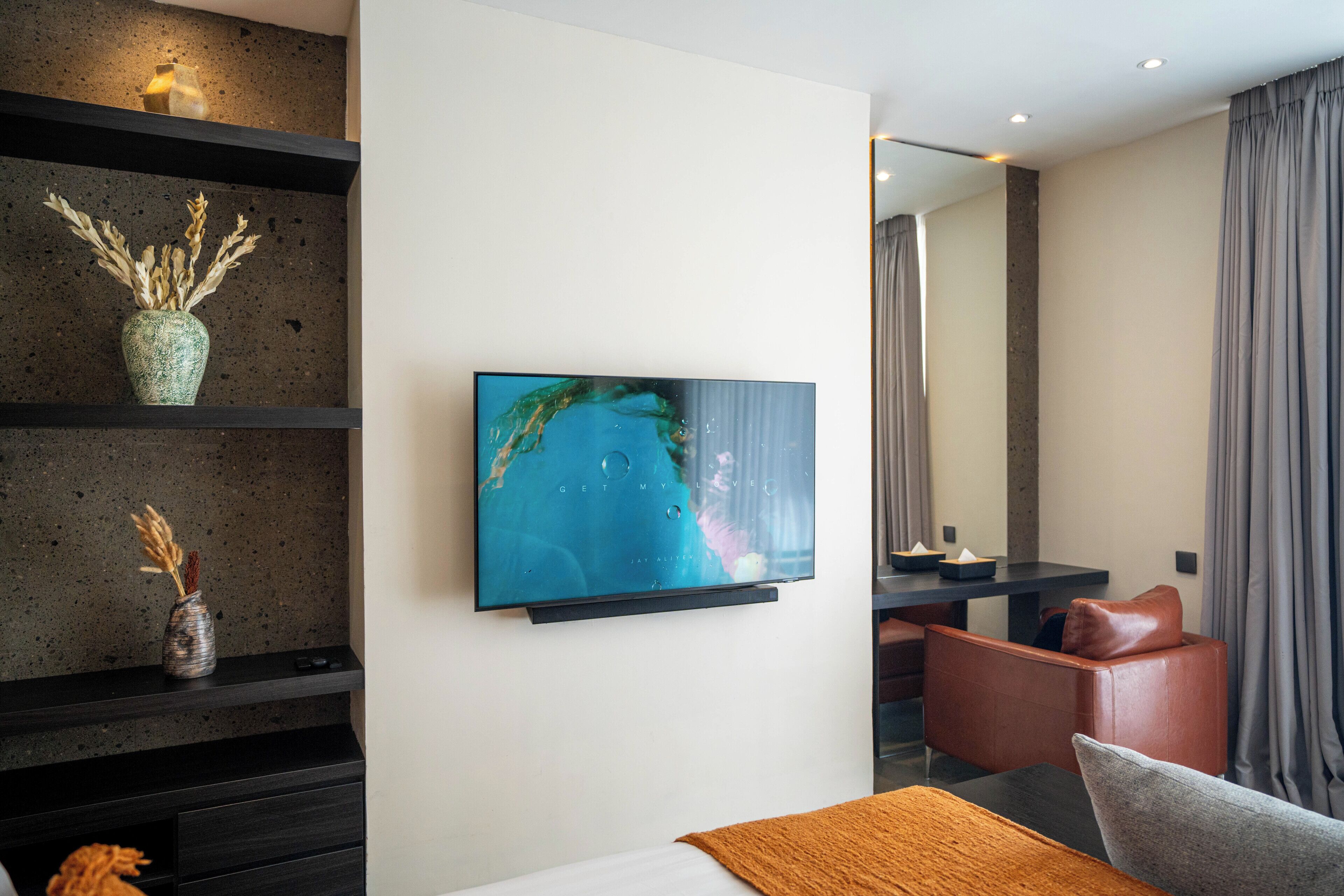 Una habitación de hotel moderna con una televisión de pantalla plana montada en la pared, un escritorio de madera oscura, un sillón de cuero marrón y una cama con una manta marrón.