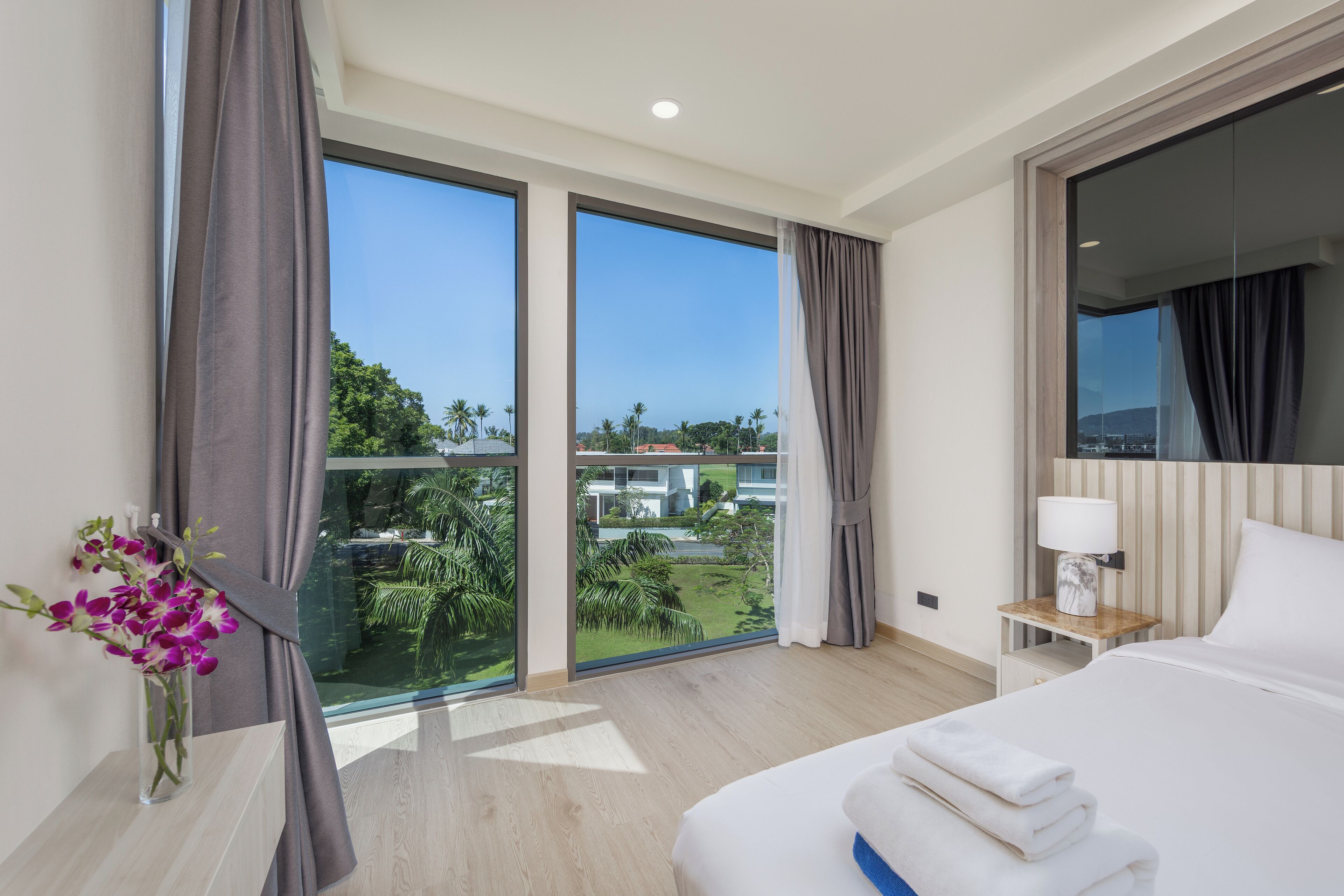Camera d'albergo con un letto grande, un comodino con una lampada, un vaso con fiori e vista su un paesaggio tropicale attraverso ampie finestre.