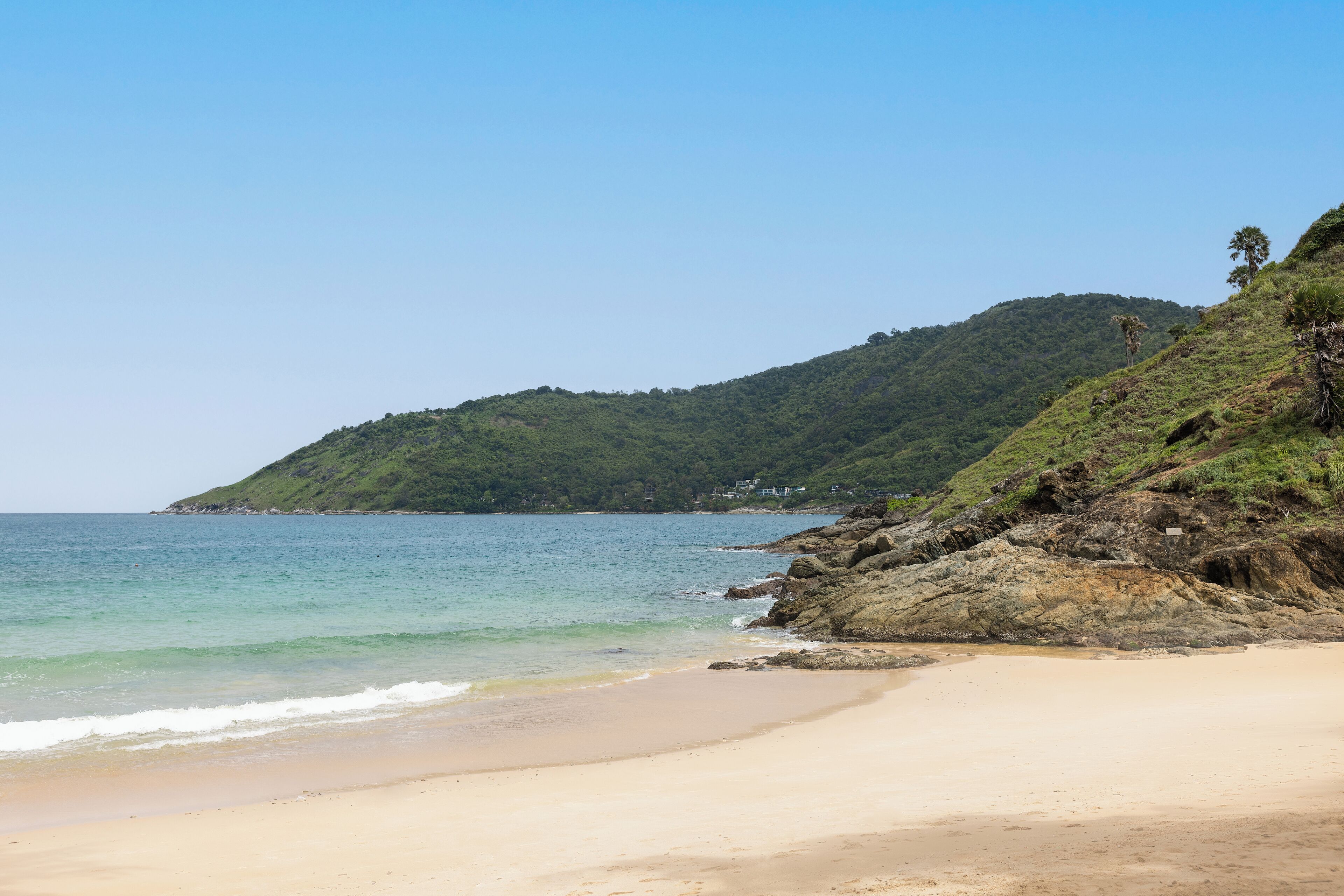 A beach with clear blue water, sandy shore, and a hill with vegetation.