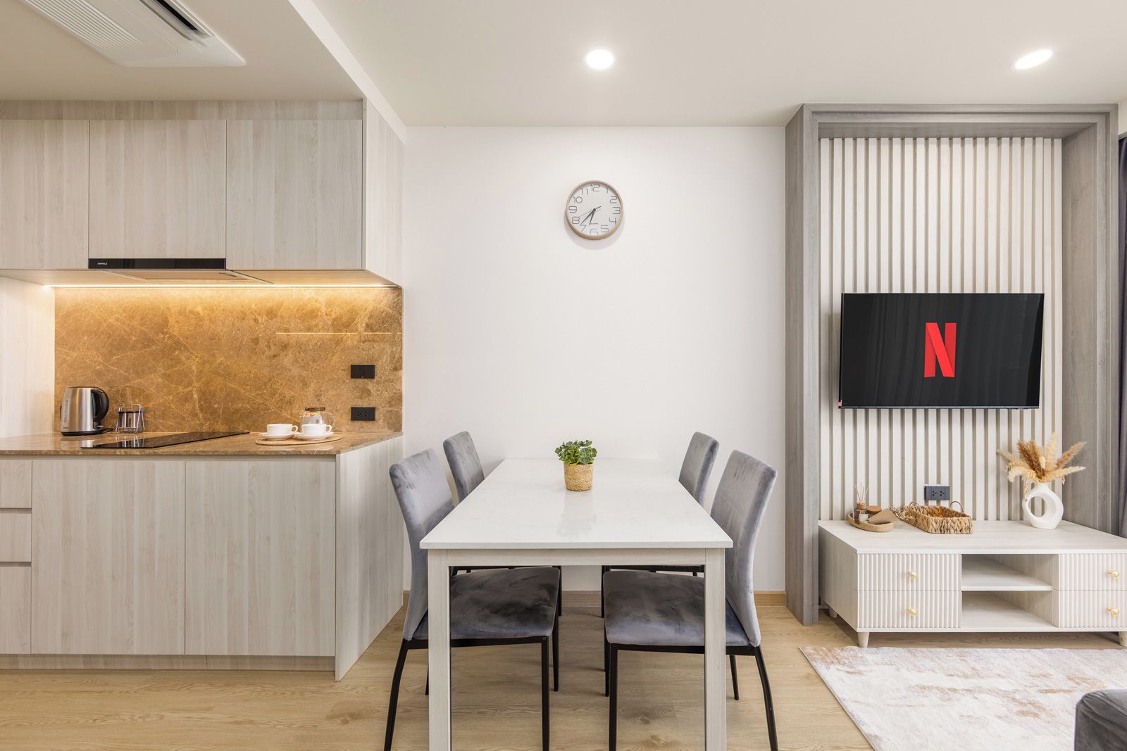 A modern kitchen with a built-in stove, a dining table with chairs, a wall-mounted clock, and a flat-screen TV displaying the Netflix logo.