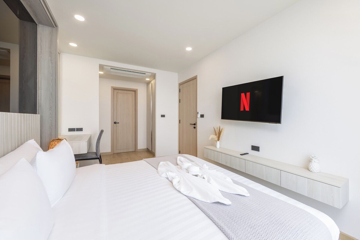 Una habitación de hotel moderna con una cama grande, un televisor de pantalla plana en la pared, una puerta de madera y una pequeña mesa con una silla.