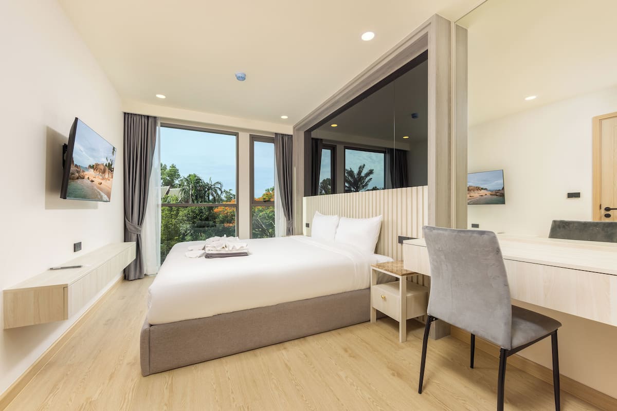 A modern hotel room with a large bed, a desk, and a chair, a TV mounted on the wall, and large windows offering a view of the outdoors.